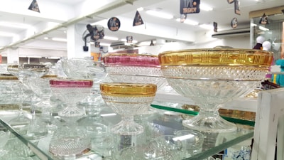 A collection of intricately designed glass bowls with a crystal-clear finish is displayed on glass shelves. The bowls have ornate patterns, with some featuring gold and pink accents on the rim. The shelves are set in a well-lit store environment, reflecting the bright lighting above.