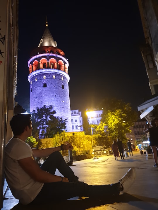 man wearing white shirt and black pants sitting on concrete floor during night time in Galata Tower Turkey