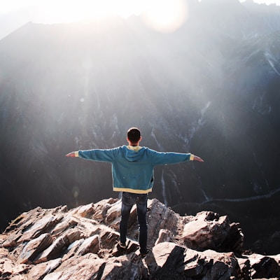 A person stands on the edge of a rocky cliff with arms outstretched, facing a vast mountainous landscape under bright sunlight. The scene conveys a sense of freedom and adventure.