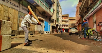 A group of people, including a person swinging a cricket bat, are in an urban street setting surrounded by shuttered shops and buildings. A person with a bicycle stands to the side, while others are visible further down the street. The street appears somewhat dilapidated.
