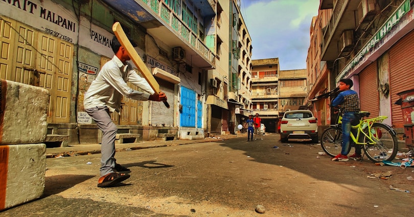A group of people, including a person swinging a cricket bat, are in an urban street setting surrounded by shuttered shops and buildings. A person with a bicycle stands to the side, while others are visible further down the street. The street appears somewhat dilapidated.