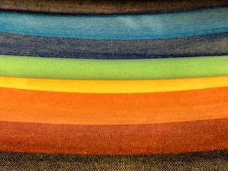 A series of fabric rolls in various bright colors are stacked horizontally. Each roll displays a distinct hue, creating a gradient effect from dark to light shades.