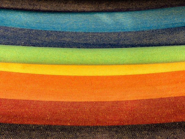 A series of fabric rolls in various bright colors are stacked horizontally. Each roll displays a distinct hue, creating a gradient effect from dark to light shades.