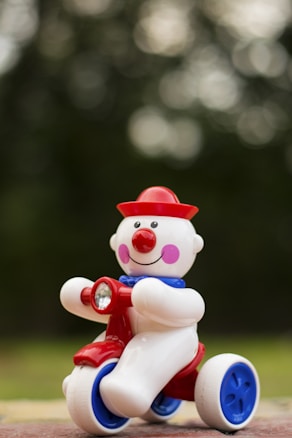 A toy clown with a round, cheerful face is riding a tricycle. The clown is characterized by its red, round nose, a bright red hat, and purple cheeks. It has a blue collar and rides a tricycle with blue and white wheels. The background is softly blurred, hinting at an outdoor setting.