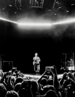 A performer stands on a stage in front of a large crowd of fans. The stage is dimly lit with a bright horizontal line of light above. Audience members hold up their phones to capture the moment.