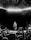 A performer stands on a stage in front of a large crowd of fans. The stage is dimly lit with a bright horizontal line of light above. Audience members hold up their phones to capture the moment.