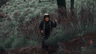 A person wearing hiking gear and a hat is smiling while walking through a lush, green forest area. The ground is uneven and covered with various types of vegetation. The scene conveys a sense of adventure and connection with nature.