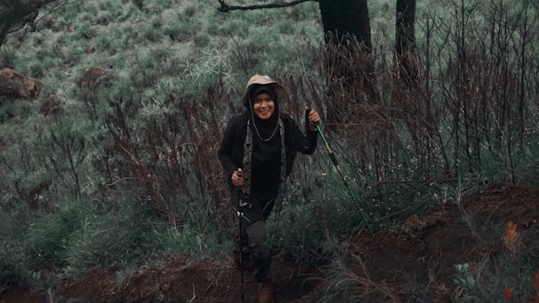 A person wearing hiking gear and a hat is smiling while walking through a lush, green forest area. The ground is uneven and covered with various types of vegetation. The scene conveys a sense of adventure and connection with nature.