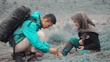 A man wearing a turquoise jacket and backpack is helping tie the shoelaces of a seated woman wearing a headscarf and dark clothing on a grassy landscape.