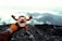 A plush toy is held up against a scenic mountain landscape, with clouds drifting over the rugged terrain. The toy is gripped in a gloved hand, suggesting a cold or adventurous environment.