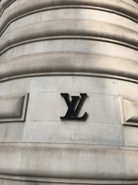 A large stone building wall featuring a prominent black metal logo with the letters 'LV' prominently mounted on its surface. The wall has a series of horizontal, rounded grooves that create a textured appearance. The structure displays an architectural style that suggests strength and luxury.
