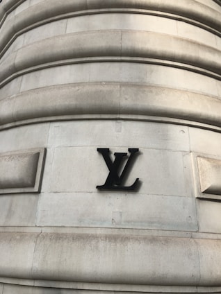 A large stone building wall featuring a prominent black metal logo with the letters 'LV' prominently mounted on its surface. The wall has a series of horizontal, rounded grooves that create a textured appearance. The structure displays an architectural style that suggests strength and luxury.