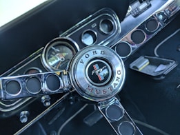 A close-up view of a classic Ford Mustang steering wheel featuring a polished metal center with the iconic horse emblem and the words 'Ford' and 'Mustang' engraved around it. The background shows part of the vehicle dashboard, including a speedometer and various controls.