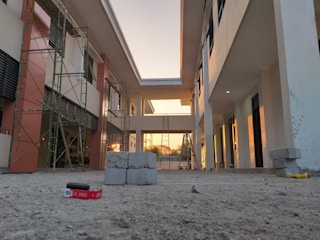 A construction site with scaffolding and partially constructed buildings. The sun is setting, casting warm tones on the walls. The ground is sandy with some construction debris and building materials scattered around.