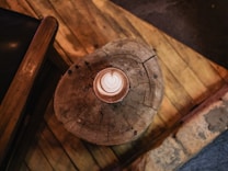A cup of coffee with latte art sits on a rustic wooden table, with visible wood grain and a circular pattern. The table appears aged and weathered, adding a cozy, vintage feel. A dark-colored chair is partially visible, contributing to the warm, inviting setting.