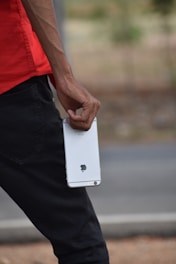 A person holding a smartphone with a visible Apple logo, wearing a red shirt and black pants. The arm is slightly veiny, and the scene appears to be outdoors with a blurred background.