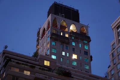 A building under construction or renovation is captured during twilight, with large arched openings at the top and illuminated windows scattered across its facade. Scaffolding and construction materials are visible, indicating ongoing work. The surrounding sky is a deep blue, typical of early evening.