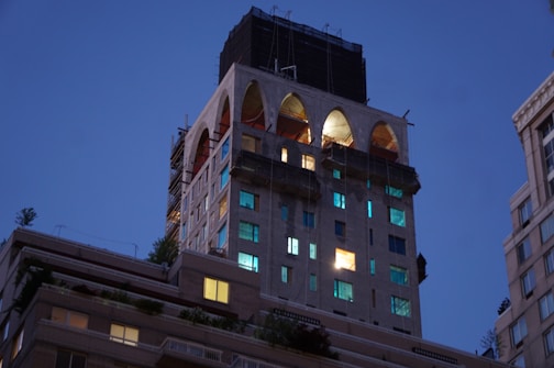 A building under construction or renovation is captured during twilight, with large arched openings at the top and illuminated windows scattered across its facade. Scaffolding and construction materials are visible, indicating ongoing work. The surrounding sky is a deep blue, typical of early evening.