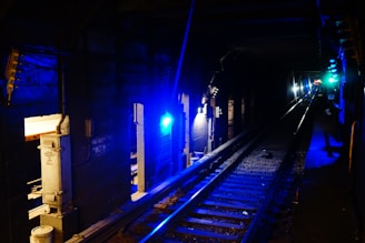 A dimly lit underground subway tunnel adorned with blue lights along the walls. The tracks extend into the darkness, and there are some electrical components and utility boxes visible along the sides.