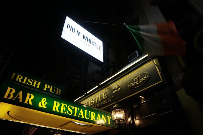 Neon signs displaying names like Pig N' Whistle and Irish Pub Bar & Restaurant are prominently featured. An Irish flag can be seen waving above the establishment, suggesting an Irish theme or affiliation.