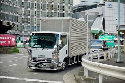 A silver Isuzu delivery truck is navigating a curved road in an urban setting. The background features a modern multi-story building with numerous windows, suggesting a city environment. A black SUV is parked nearby, and a pedestrian is visible. Street signs and barrier rails are present, giving a sense of a well-trafficked area.