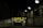 A dimly lit entrance to a parking garage with a prominent yellow and black sign reading 'PARK OPEN 24 hours.' The sign provides additional cautionary details about clearance and vehicle restrictions. The setting is enclosed by brick walls with several overhead lights illuminating parts of the area.
