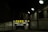 A dimly lit entrance to a parking garage with a prominent yellow and black sign reading 'PARK OPEN 24 hours.' The sign provides additional cautionary details about clearance and vehicle restrictions. The setting is enclosed by brick walls with several overhead lights illuminating parts of the area.