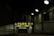 A dimly lit entrance to a parking garage with a prominent yellow and black sign reading 'PARK OPEN 24 hours.' The sign provides additional cautionary details about clearance and vehicle restrictions. The setting is enclosed by brick walls with several overhead lights illuminating parts of the area.