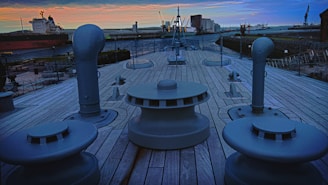 The deck of a ship with large bollards and vents is shown, surrounded by a shipyard. The sky is filled with orange and blue hues at sunset. Ships and cranes are visible in the background.