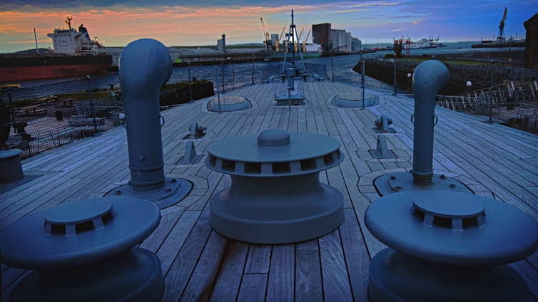 The deck of a ship with large bollards and vents is shown, surrounded by a shipyard. The sky is filled with orange and blue hues at sunset. Ships and cranes are visible in the background.