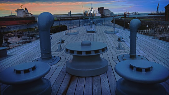 The deck of a ship with large bollards and vents is shown, surrounded by a shipyard. The sky is filled with orange and blue hues at sunset. Ships and cranes are visible in the background.