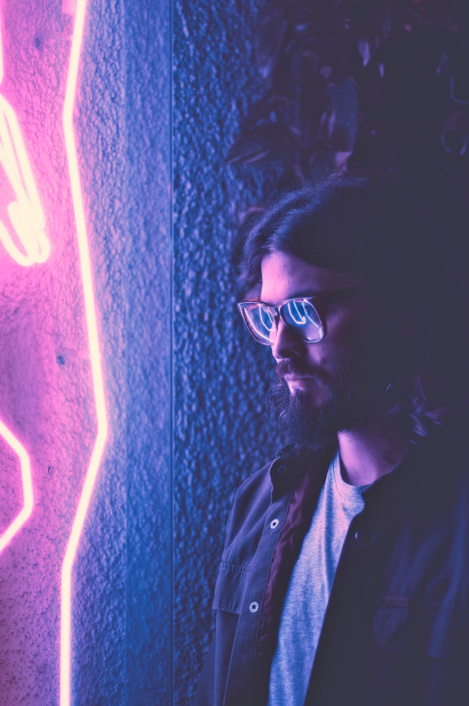 The image shows a man with long hair and glasses standing against a blue wall, wearing a gray jacket. The man is looking off to the side, possibly observing something or someone outside the frame. A neon sign in shades of pink and purple is visible on the right side of the photo, adding an element of modernity and vibrancy to the scene.

The image also contains text that reads "Be as descriptive as possible" and "Image description".
