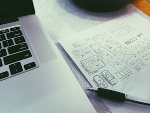 A close-up view of a laptop keyboard and a sheet of paper with various hand-drawn sketches. The sketches include wireframes, geometric shapes, and typography. A black pen is placed diagonally on the paper.