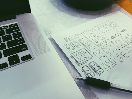 A close-up view of a laptop keyboard and a sheet of paper with various hand-drawn sketches. The sketches include wireframes, geometric shapes, and typography. A black pen is placed diagonally on the paper.