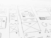 A detailed sketch of web design layouts, featuring boxes, arrows, and various shapes representing components of a webpage. The designs include placeholders for images and text, with lines and symbols indicating structure.