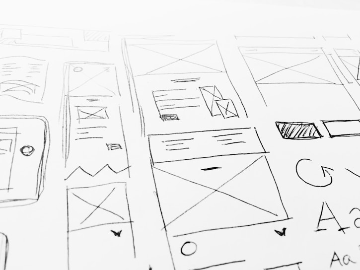 A detailed sketch of web design layouts, featuring boxes, arrows, and various shapes representing components of a webpage. The designs include placeholders for images and text, with lines and symbols indicating structure.