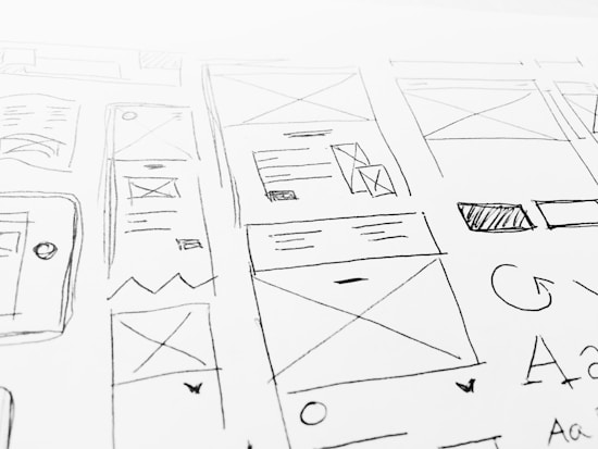 A detailed sketch of web design layouts, featuring boxes, arrows, and various shapes representing components of a webpage. The designs include placeholders for images and text, with lines and symbols indicating structure.