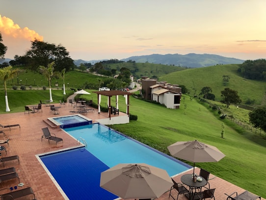 A serene outdoor setting with a swimming pool surrounded by lounge chairs and tables with umbrellas on a paved patio. The backdrop features lush green hills under a clear sky with clouds, and a collection of modern buildings nestled on the hillside. The landscape is rich with green grass and interspersed trees, giving a sense of relaxation and tranquility.