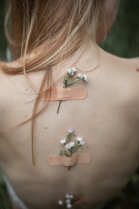 A close-up of a person's bare back with small flowers inserted into skin-colored adhesive bandages. The flowers are delicate and white, giving a natural and serene feel. The person's long hair drapes softly over the shoulder.