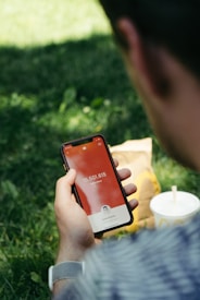 A person is holding a smartphone displaying a subscriber count on the screen, with the number 15,501,615 visible alongside a small profile picture. In the background, a fast-food drink cup and a paper bag sit on the grass, suggesting a casual outdoor environment.