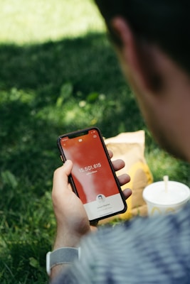 A person is holding a smartphone displaying a subscriber count on the screen, with the number 15,501,615 visible alongside a small profile picture. In the background, a fast-food drink cup and a paper bag sit on the grass, suggesting a casual outdoor environment.