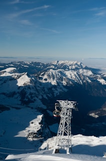 A stunning mountainous landscape with snow-covered peaks under a clear blue sky. A cable car is suspended from a towering metallic pylon, offering transport across the picturesque views. The mountains stretch into the distance, with various shadows and highlights creating depth and contrast.