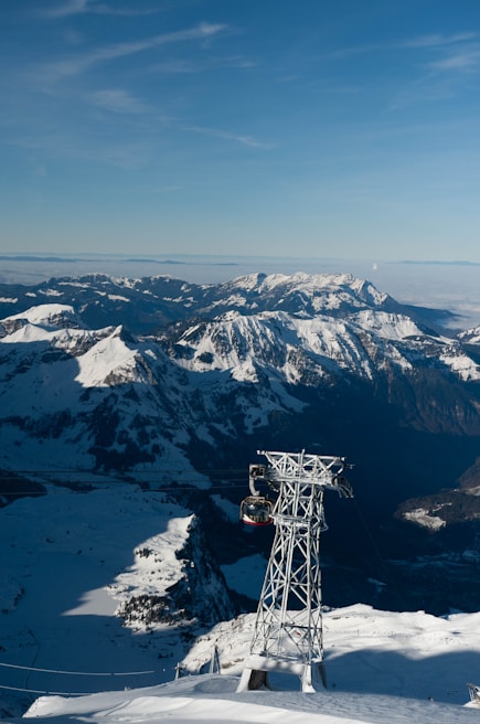 A stunning mountainous landscape with snow-covered peaks under a clear blue sky. A cable car is suspended from a towering metallic pylon, offering transport across the picturesque views. The mountains stretch into the distance, with various shadows and highlights creating depth and contrast.