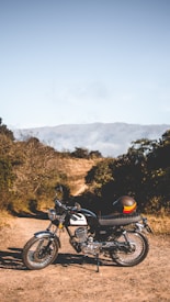 A classic motorcycle is parked on a dirt path amidst a rugged landscape with sparse vegetation. The background features mountainous terrain under a clear blue sky. Next to the motorcycle is a helmet with colorful stripes.