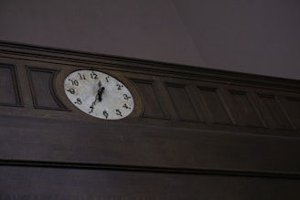 An analog wall clock is mounted on dark wood paneling. The clock features a white face with black numerals and hands, set in a traditional style.