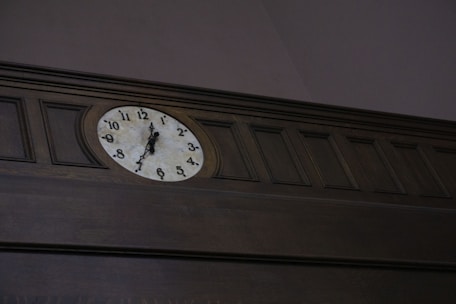 An analog wall clock is mounted on dark wood paneling. The clock features a white face with black numerals and hands, set in a traditional style.
