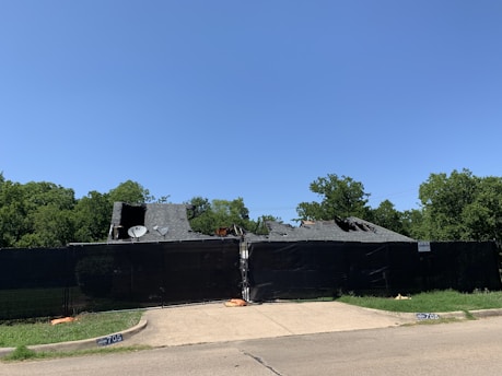 A damaged house is surrounded by a tall black fence with visible tarps and a collapsed roof. The foreground shows a concrete driveway bordered by patches of green grass. In the background, lush green trees stand against a clear blue sky.