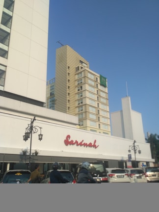 A large white building with the name 'Sarinah' in red letters is visible in the foreground. Behind it, there are several tall modern buildings with numerous windows and a clear blue sky in the background. On the street below, there are cars and motorcycles with people wearing helmets.