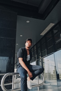A person wearing a black shirt and dark pants sits on a metal railing, looking thoughtfully into the distance. The background includes a modern architectural setting with large glass windows and a dark ceiling.