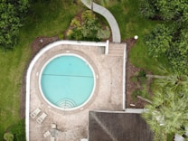 An aerial view of a circular swimming pool surrounded by a stone patio. There are several lounge chairs and a round table near the pool. The area is bordered by well-maintained green grass and various plants, with a pathway leading away from the pool area.
